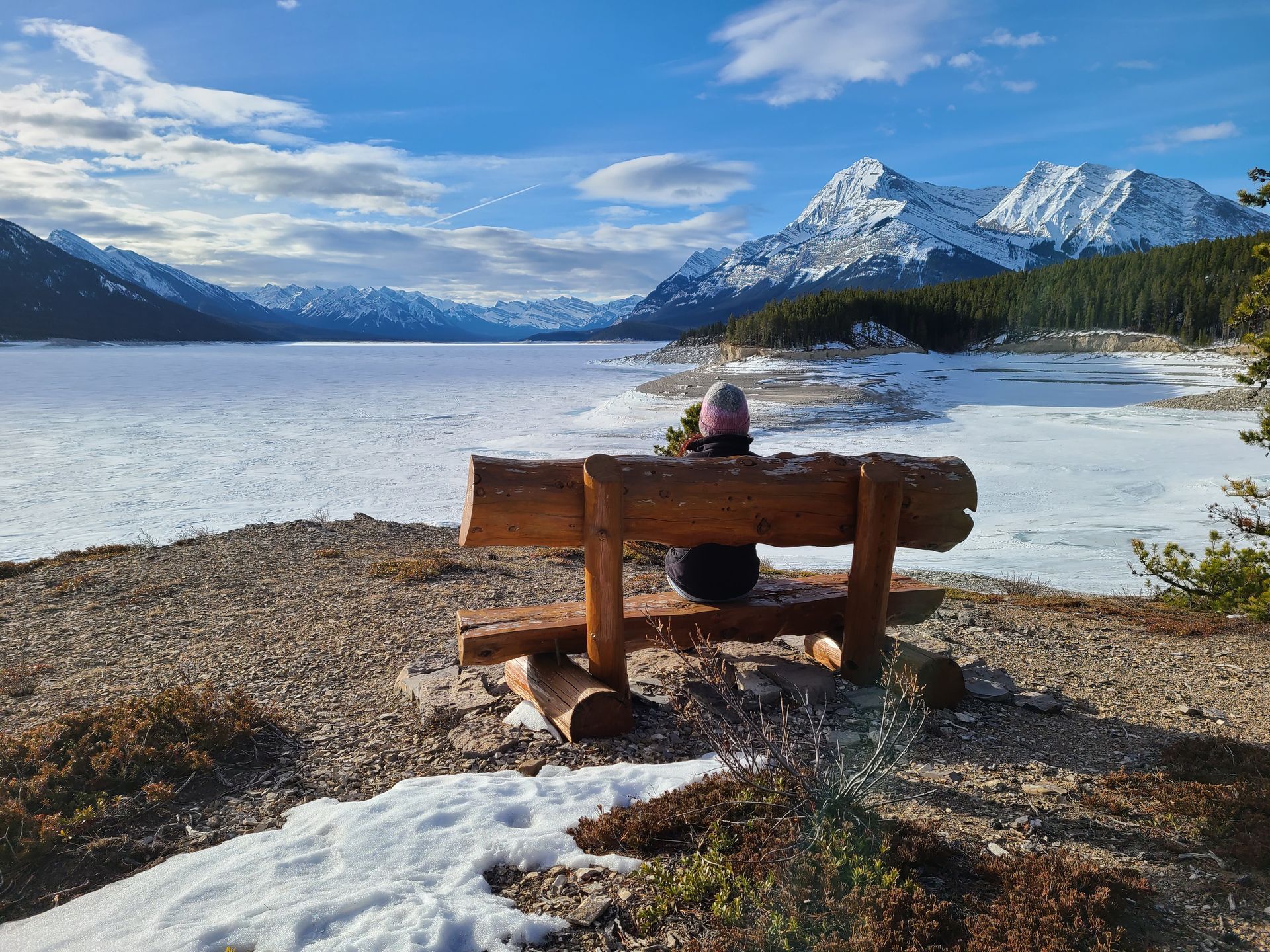 A person is sitting on a bench overlooking a lake with mountains in the background.