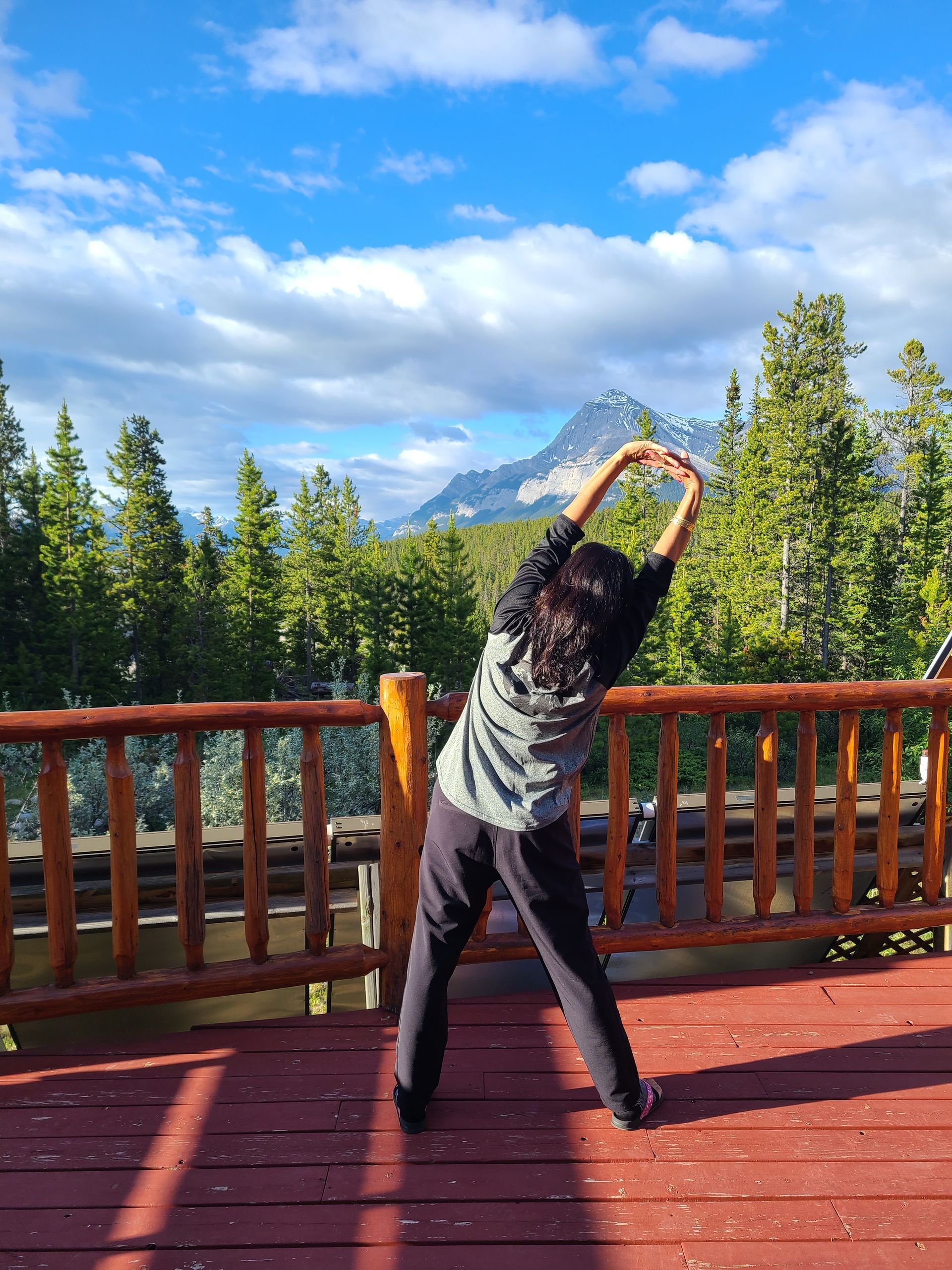 A woman is stretching her arms on a deck with a mountain in the background.