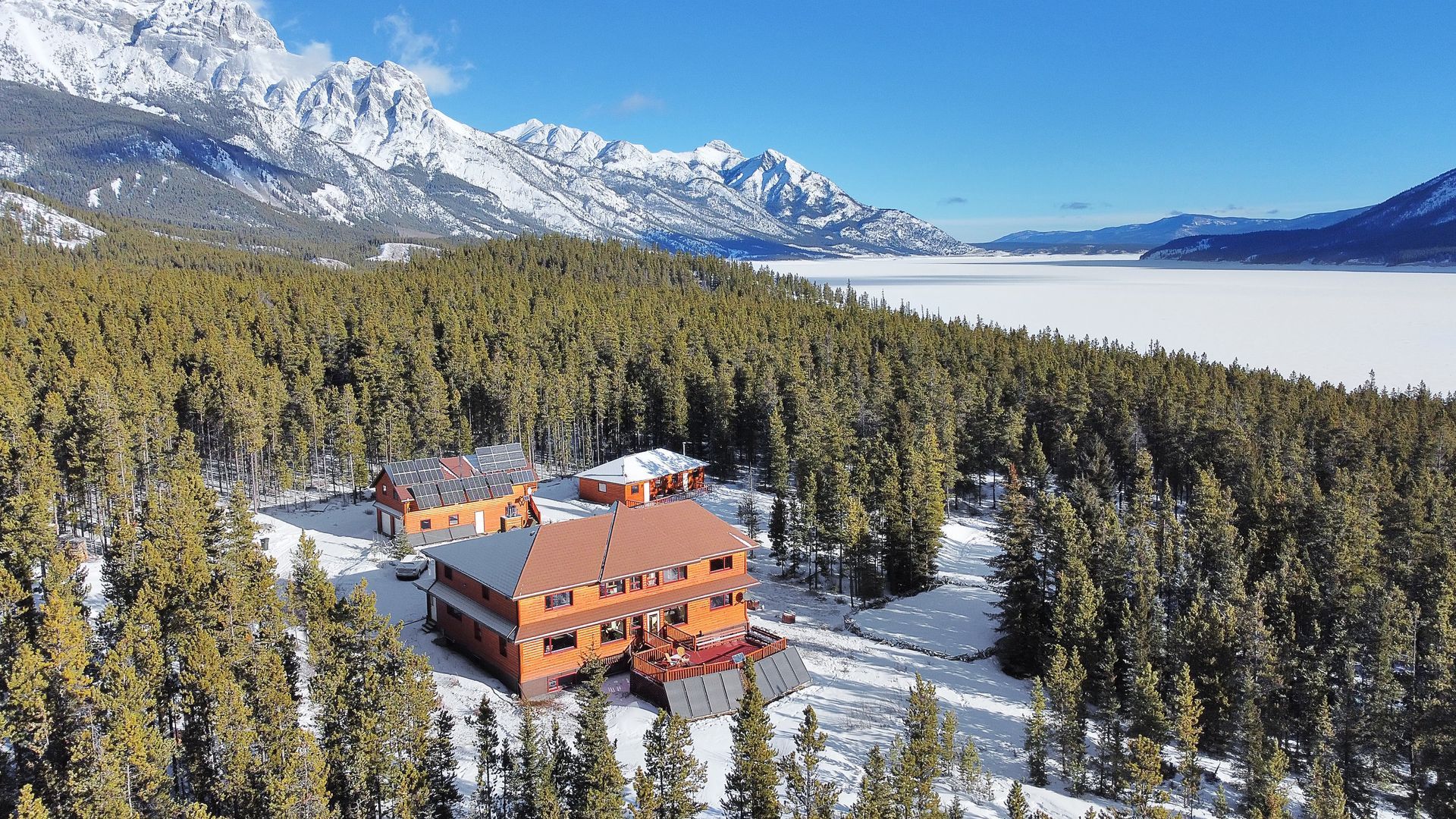 Snow-covered lodge nestled in evergreen trees with snowy mountains and frozen lake in the background.
