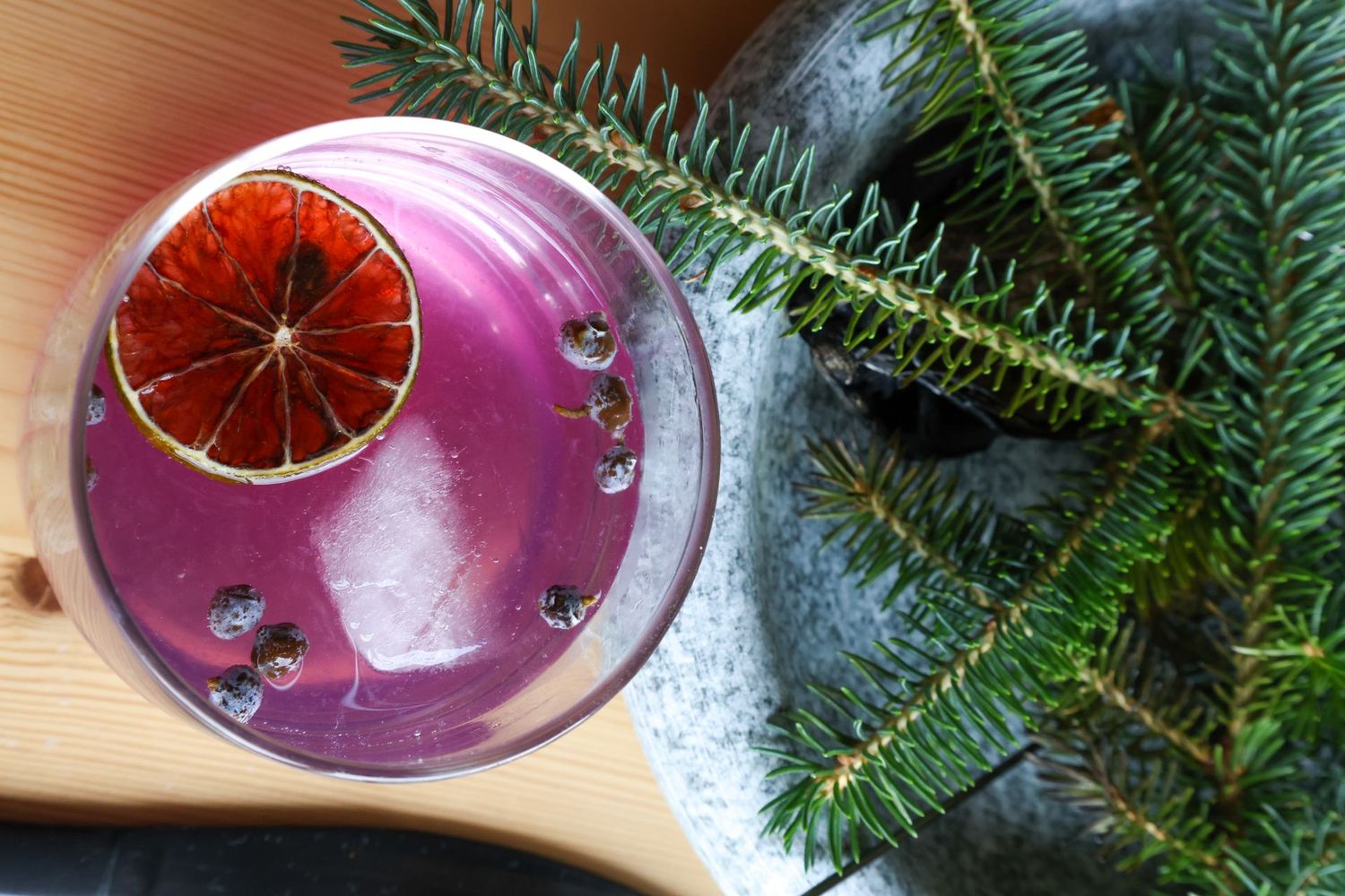 A purple cocktail with a slice of orange on top is on a table next to a christmas tree.