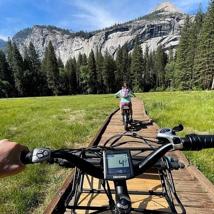 Person rides an electric bike on a boardwalk path through a meadow in front of a mountain.