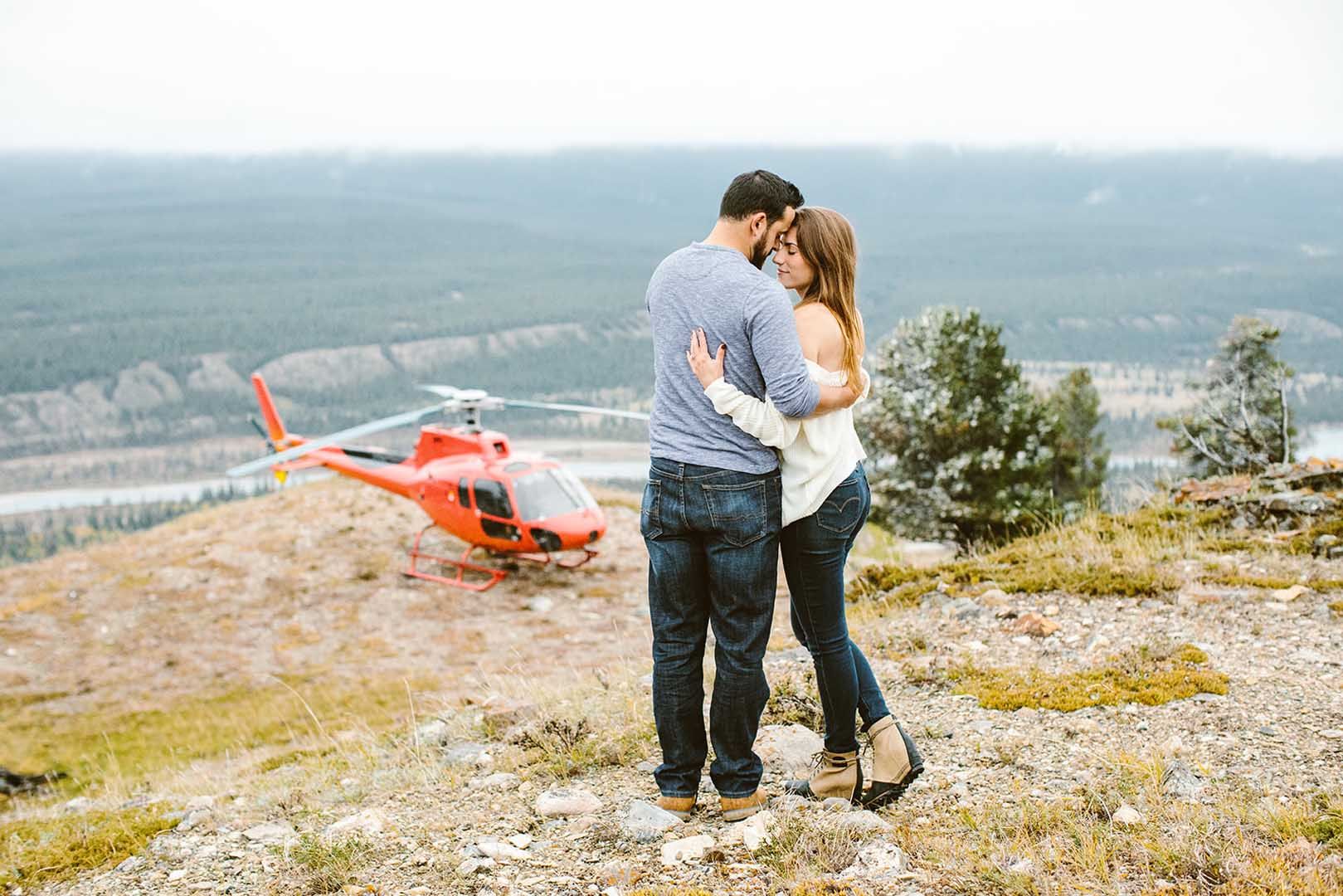 A man and a woman are hugging on top of a hill in front of a helicopter.