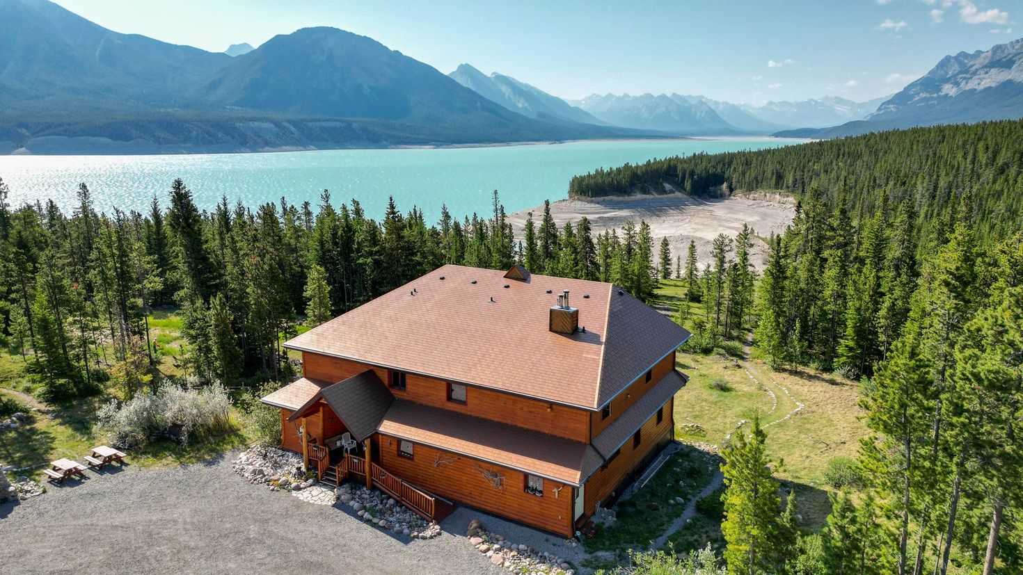 An aerial view of a log cabin surrounded by trees and mountains with a lake in the background.