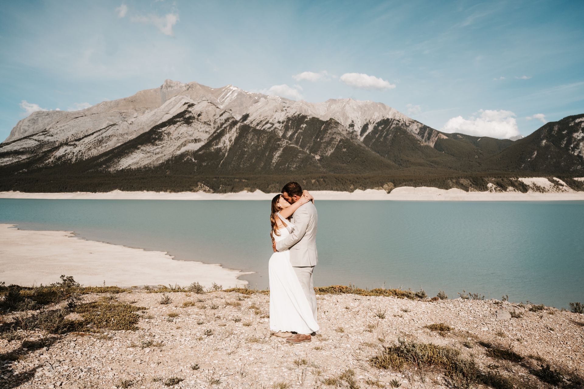 A bride and groom are hugging in front of a lake with mountains in the background.