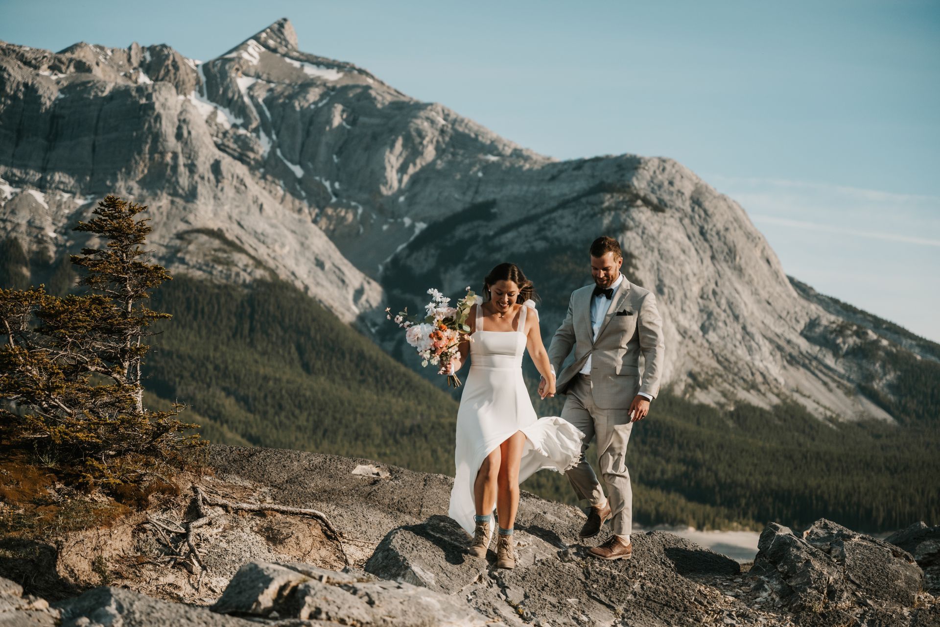 A bride and groom are standing on top of a mountain holding hands.