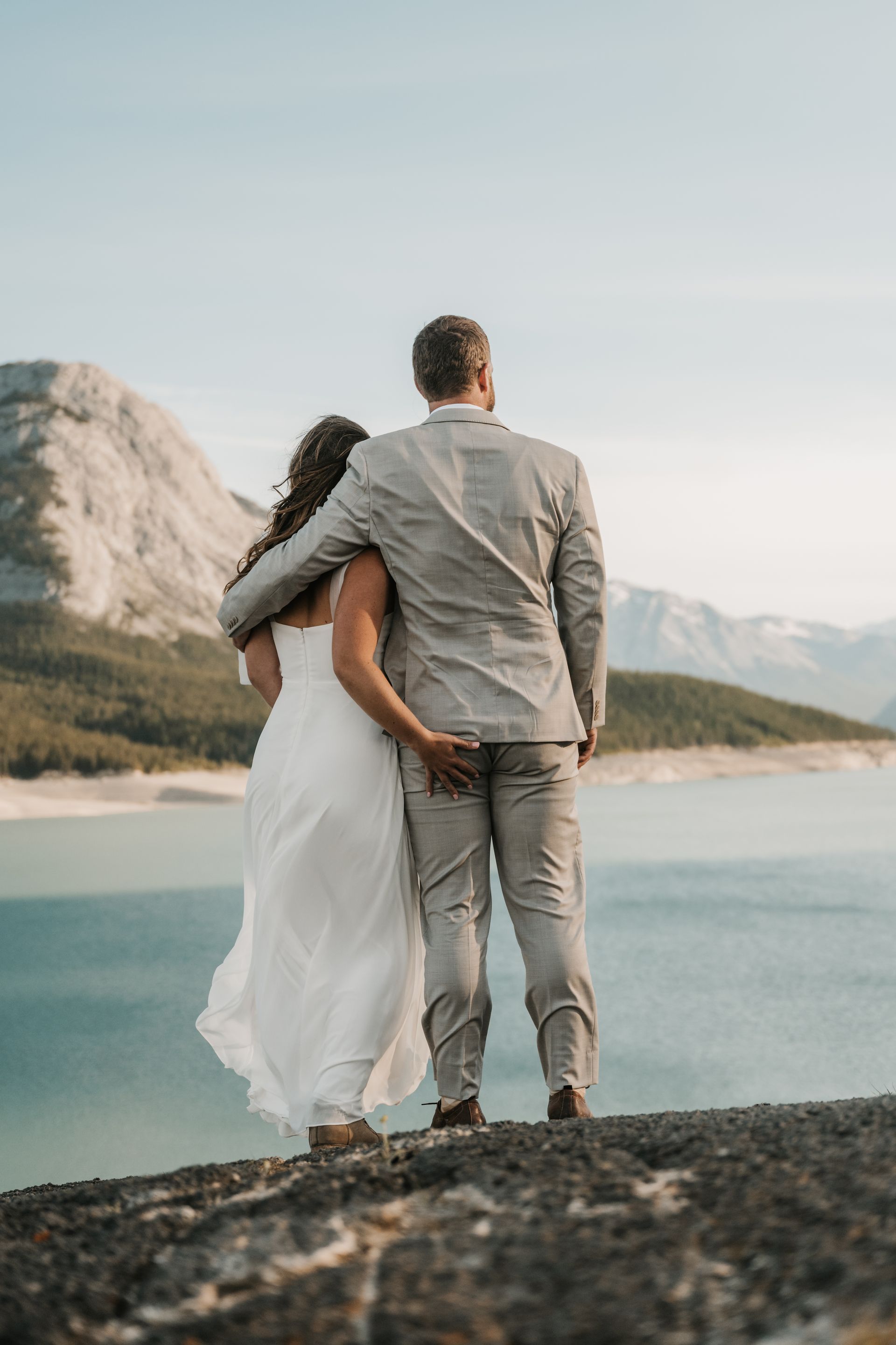 A bride and groom are standing on top of a rock overlooking a lake.