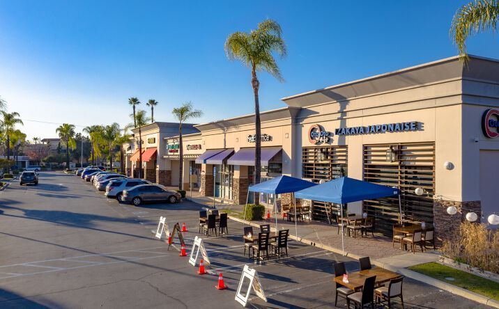 Street with shops and outdoor seating; palm trees and cars are visible.
