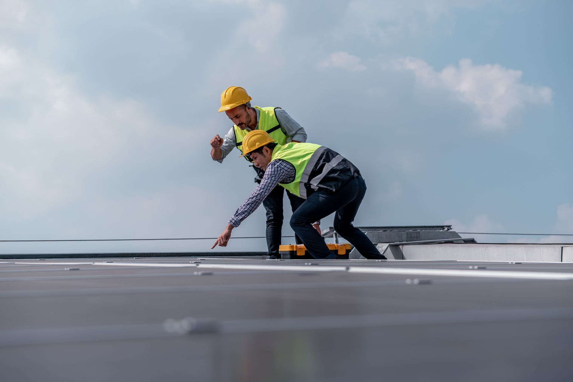 Man in hard hat and vest using a drill to install gutter on a house under a clear blue sky.