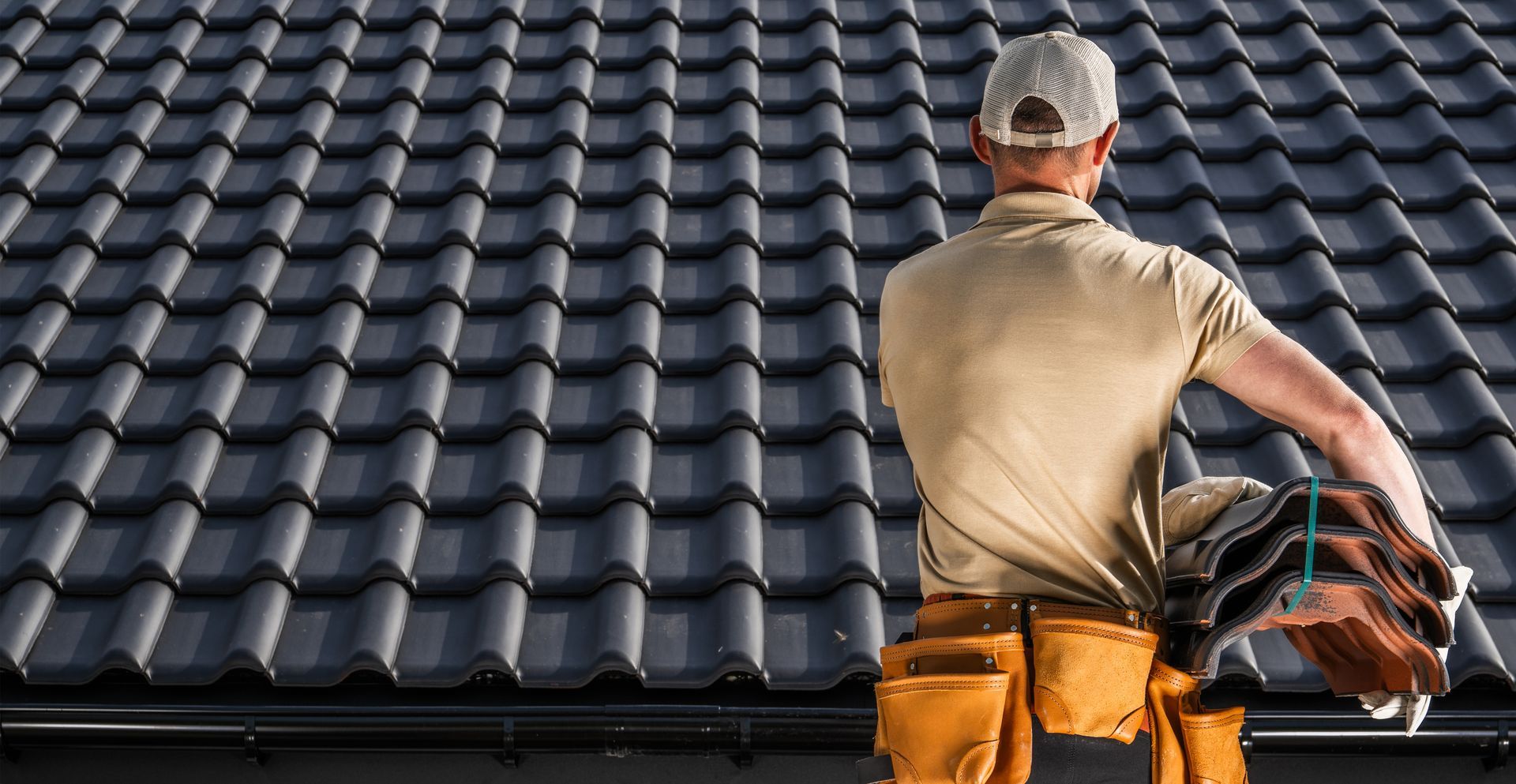 Roofer holding roofing tiles on a dark-tiled roof, wearing a tool belt and cap.