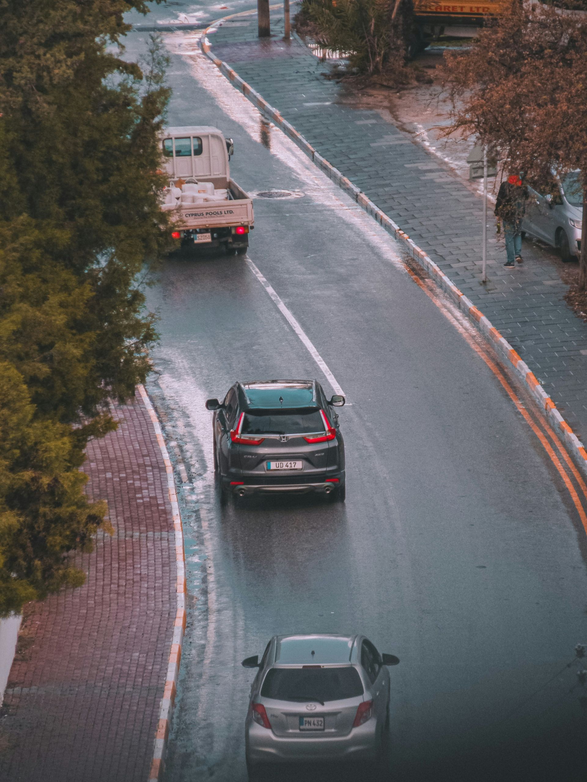 A high-angle view of a gray car following a dark SUV and a light pickup truck on a wet city street lined with trees.