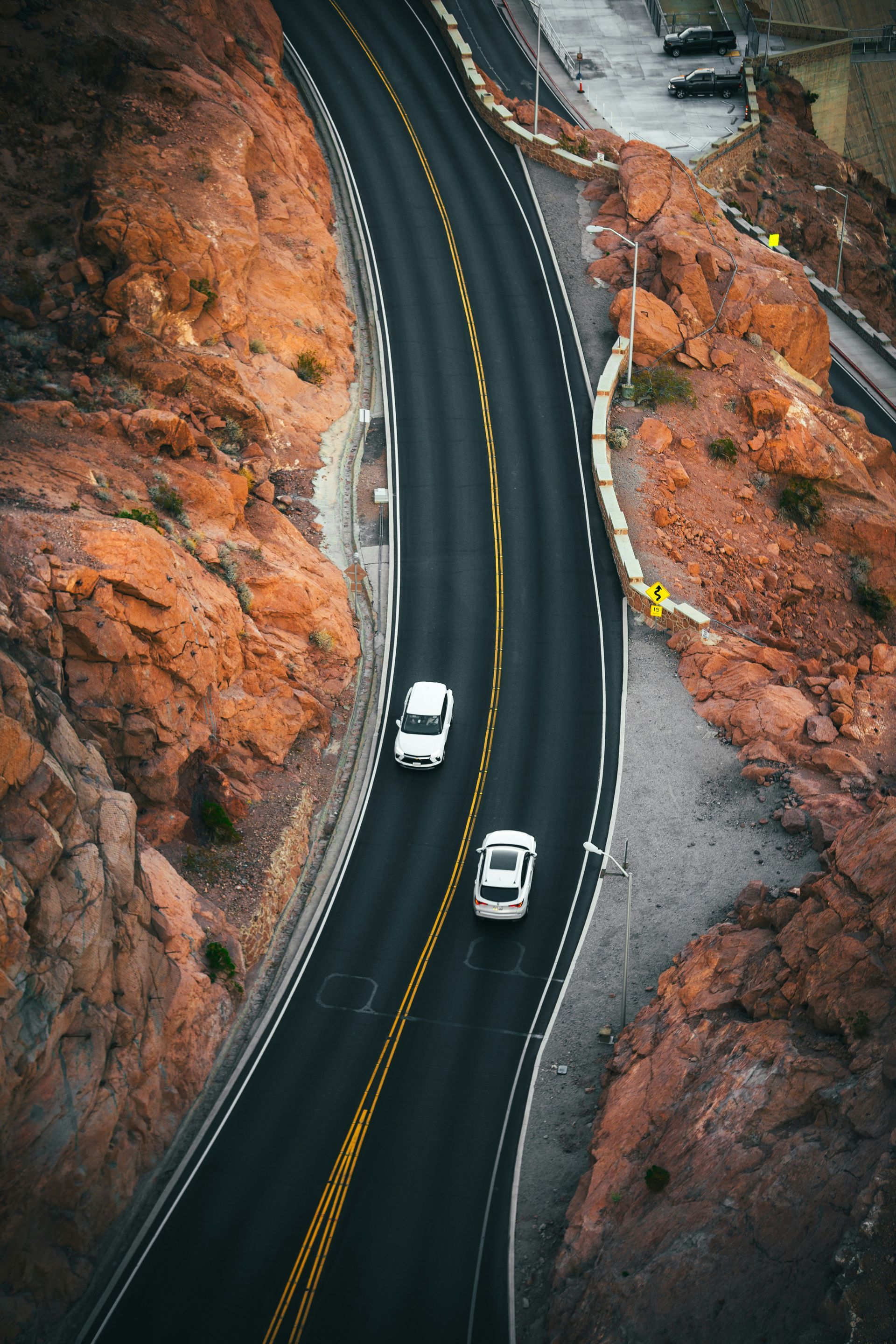 A vertical, high-angle shot of two white cars driving along a curved mountain road flanked by rugged orange rock faces.