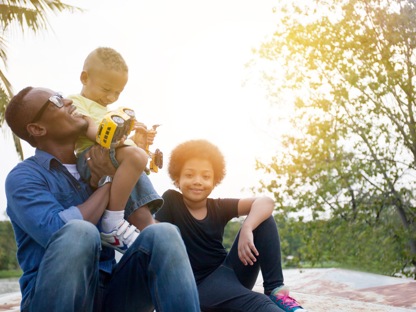 father and sons smiling together in the park playing with toy trucks