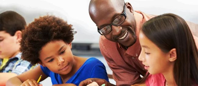Teacher helping students with a classroom activity at a table, smiling and leaning in close