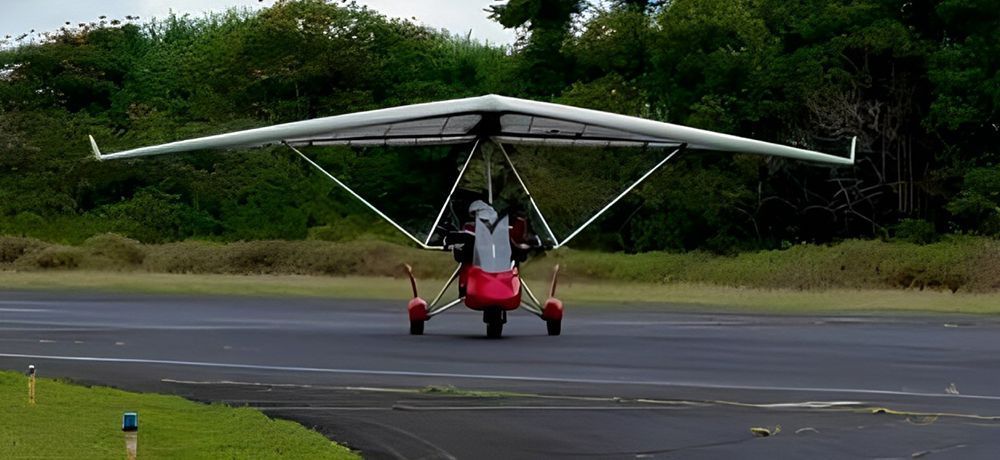 A trike aircraft with a red body and a large white wing sits on a runway, with a green backdrop of trees.