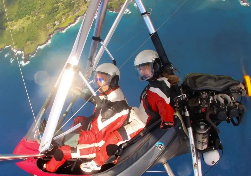 Two people in helmets fly in a powered hang glider over a coastline with blue water and green land.