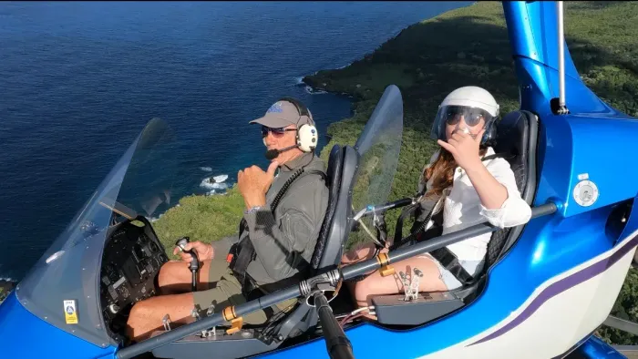 Two people in a blue helicopter flying over a coastline, pilot with a thumbs up, passenger with a shaka hand gesture.
