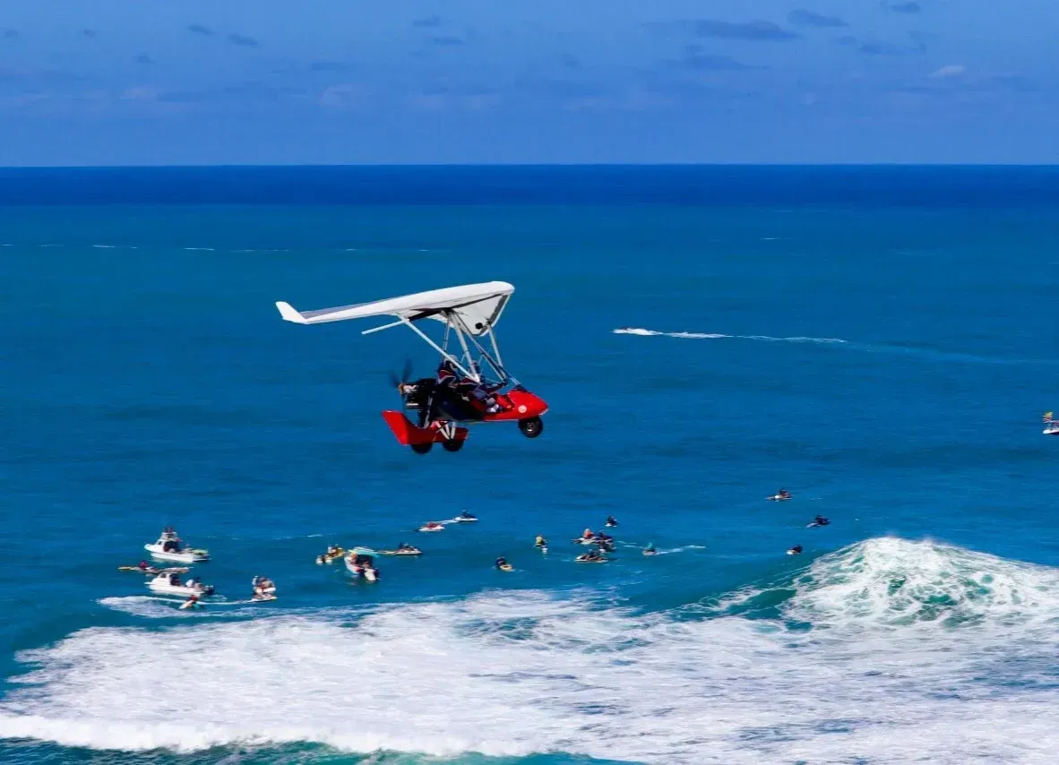 A red powered hang glider soars over the blue ocean.