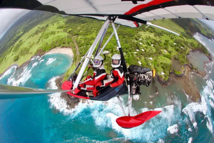 Hang glider soaring over a vibrant coastline, featuring two people in red suits, with a red wing, blue ocean and green hills.