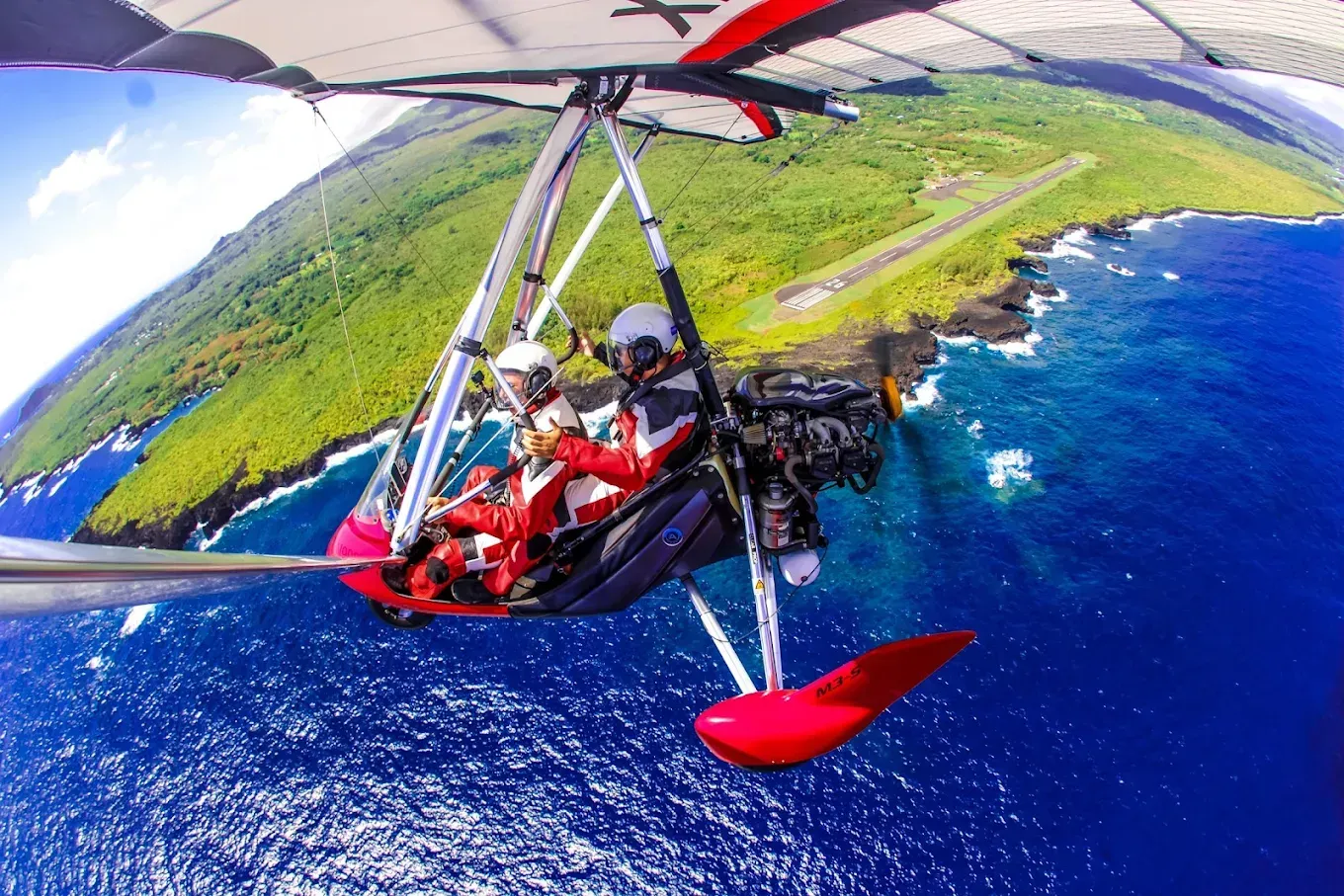Two people flying in a red and white powered hang glider over a tropical coastline.