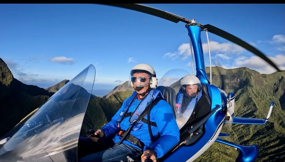 A man in a blue jacket and white helmet flies a blue helicopter with a passenger, mountains in the background.
