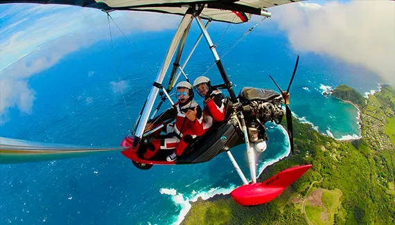 Two people fly in a red powered hang glider over a turquoise ocean and lush green coast.