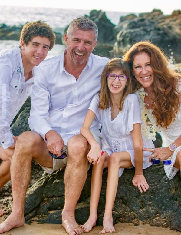 Family of four smiling on a beach, wearing white clothing. A young girl sits in front, appearing thin, wearing glasses.
