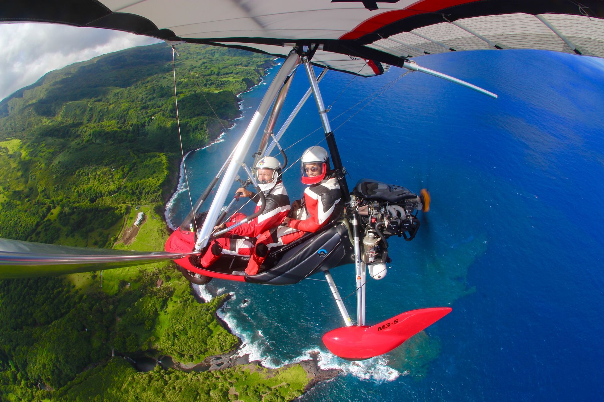 Two people in a red powered paraglider fly over a blue ocean and green coastline.