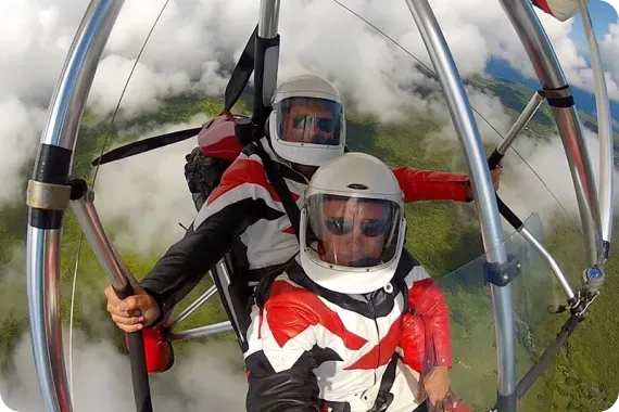 Two people in a hang glider, wearing helmets and red/black flight suits, soaring above clouds and green landscape.