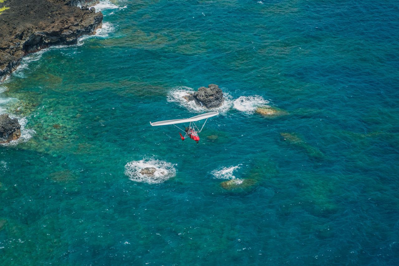 A powered paraglider flies low over the ocean, near rocky shorelines. Turquoise water surrounds it.