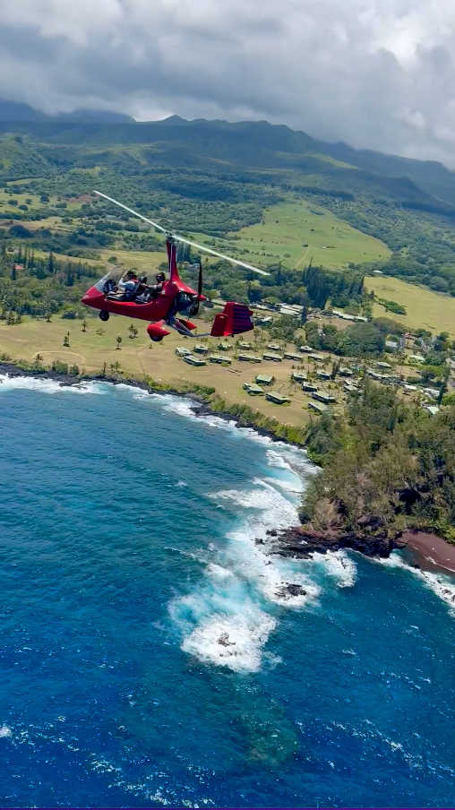 Red helicopter flying over a blue ocean and rocky coastline with a village nestled into lush green hills.