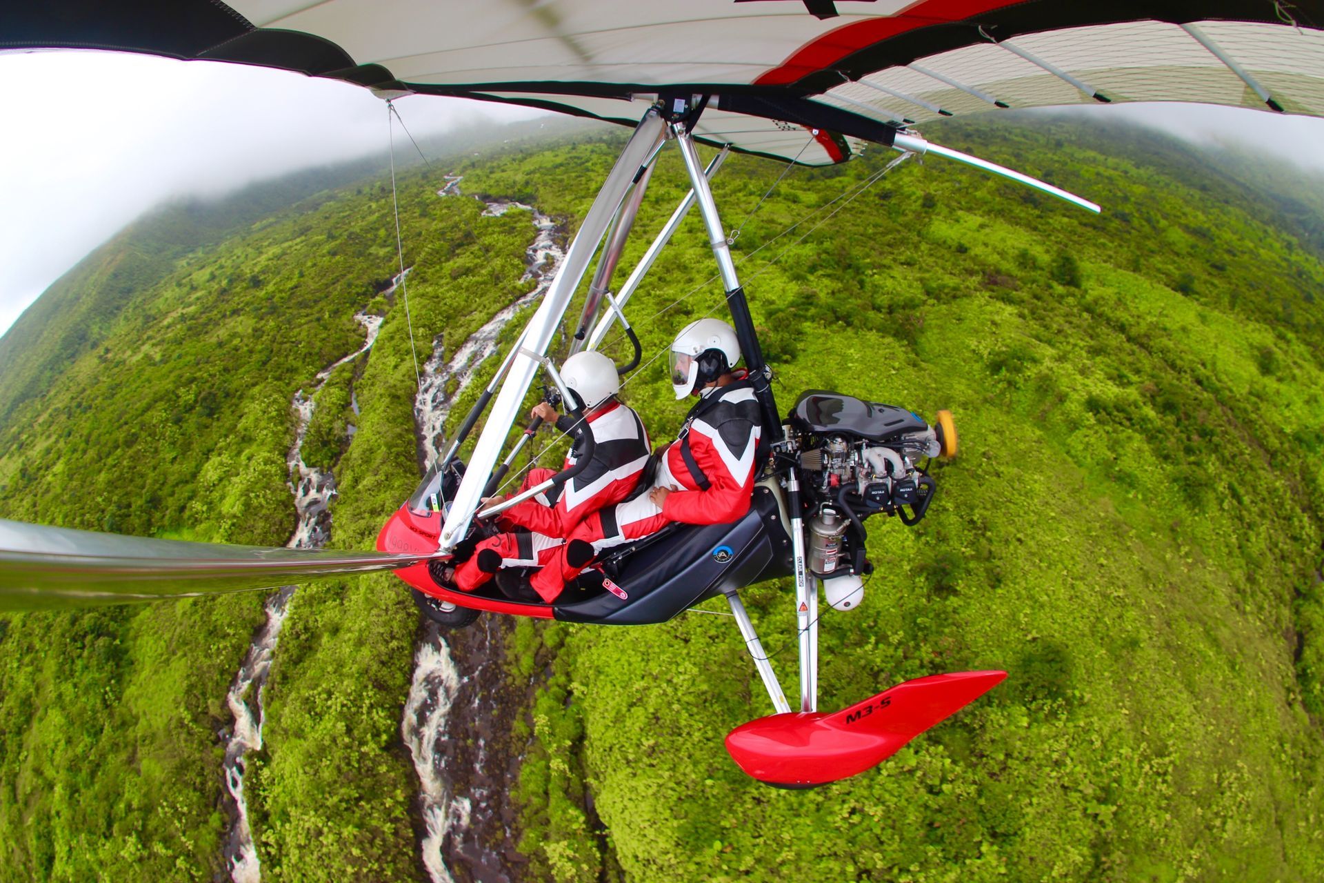 Hang glider with two people soaring over a lush, green mountain range. The glider is red and silver, and the sky is overcast.