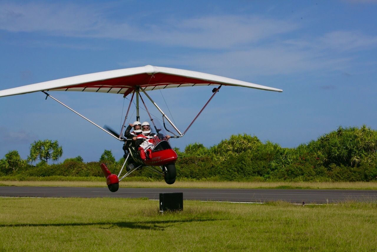 A red and white ultralight aircraft taking off from a grassy airstrip under a blue sky.