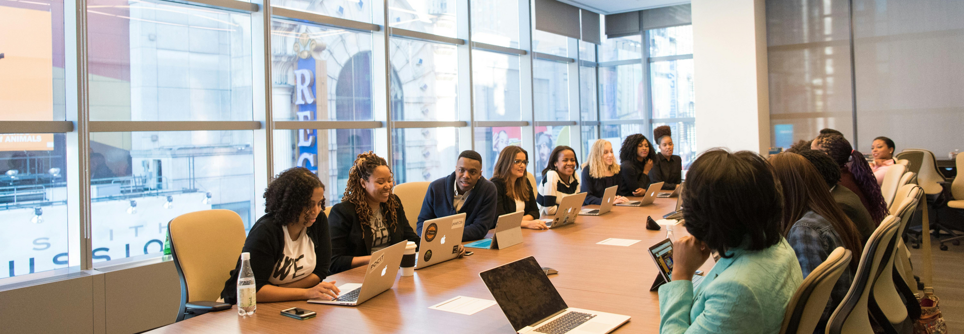 A group of diverse people sit around a large table in a bright office meeting room.