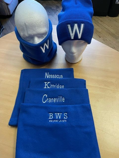Blue knit hat and neck gaiters on display, featuring embroidered school names and the BWS Bobcats logo on a wooden table.