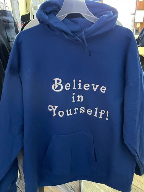 A blue hooded sweatshirt hanging on a hanger, featuring the white embroidered text 