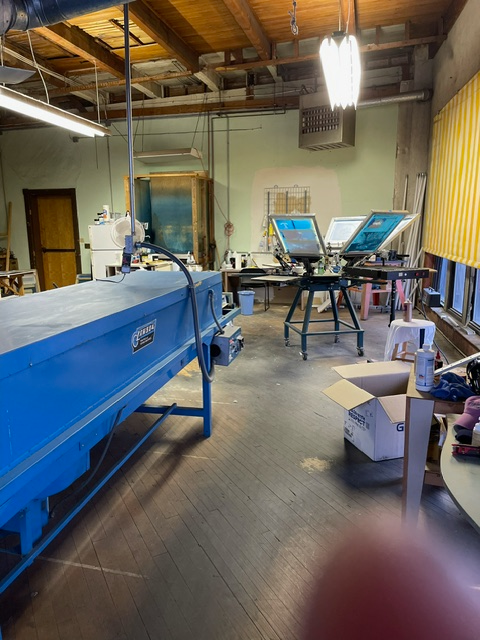 A screen printing studio featuring a blue conveyor dryer in the foreground and a multi-station printing press behind it.