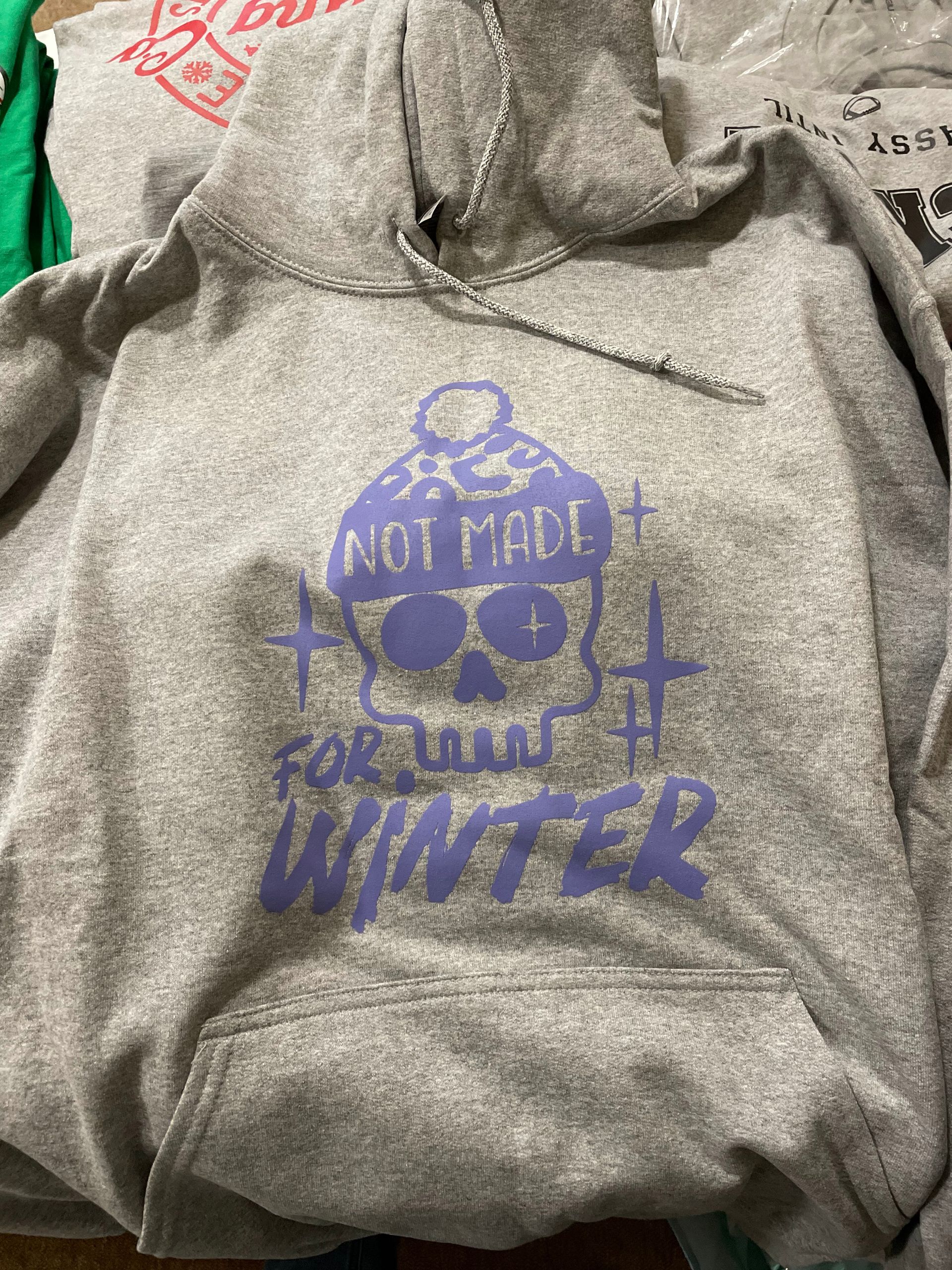 A heather gray hoodie featuring a lavender graphic of a skull wearing a beanie, with the text 