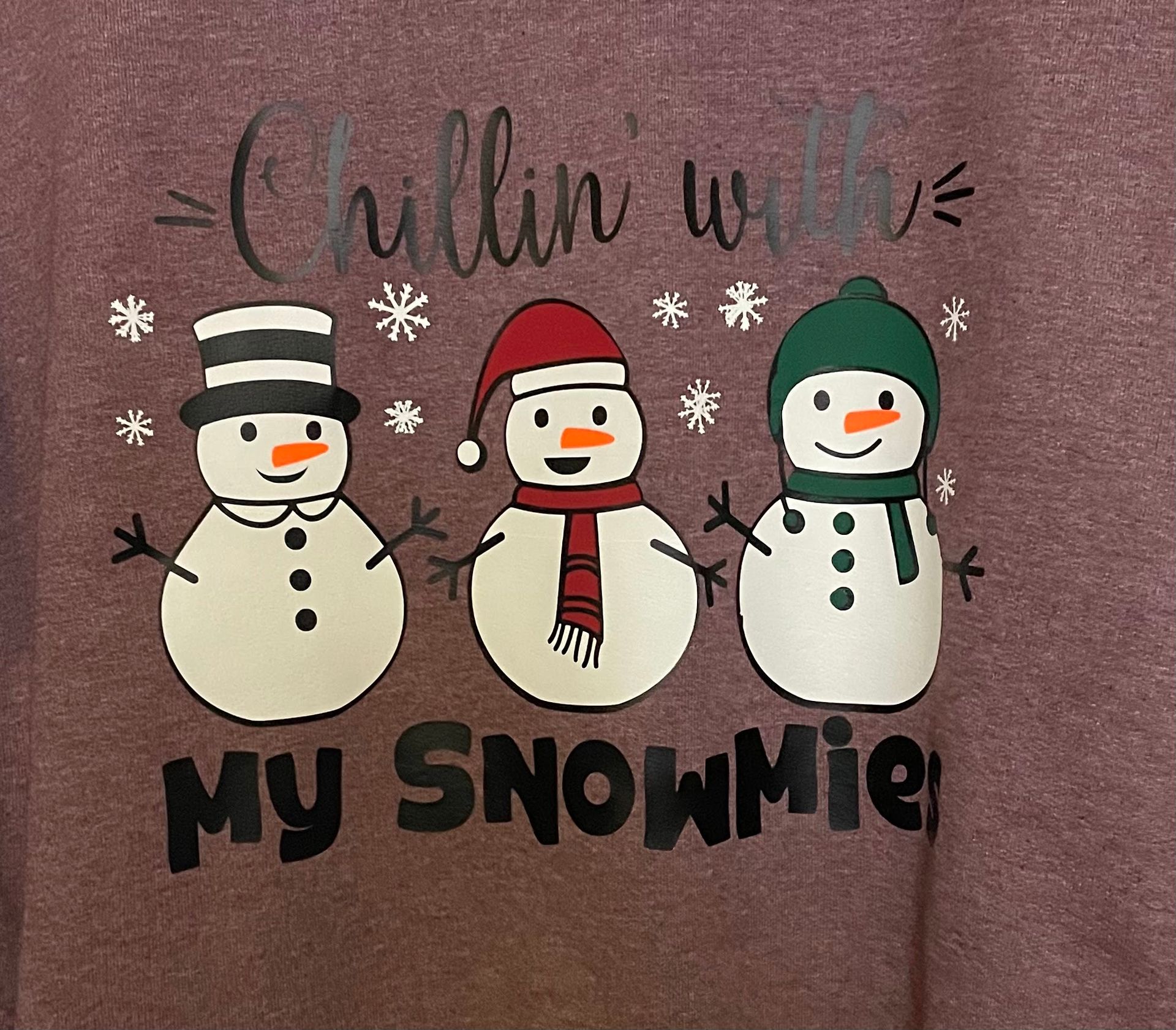 Three snowmen in hats and scarves on a maroon background with snowflakes and the text 