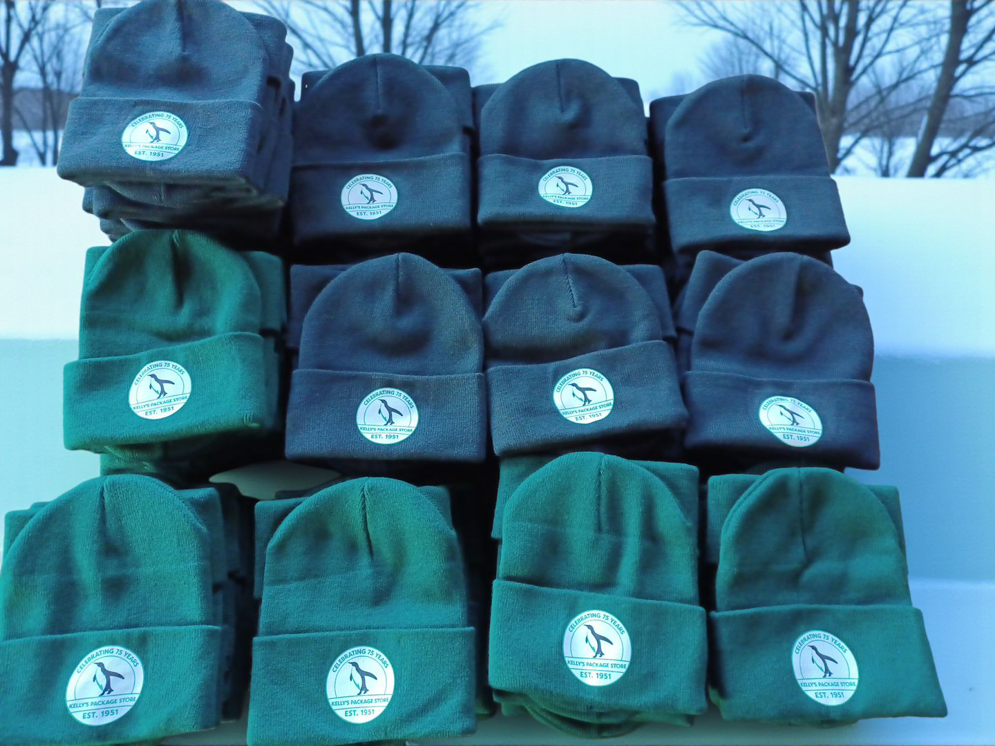 A stack of dark green and black winter beanies, each featuring a circular patch with a logo, arranged in a grid.