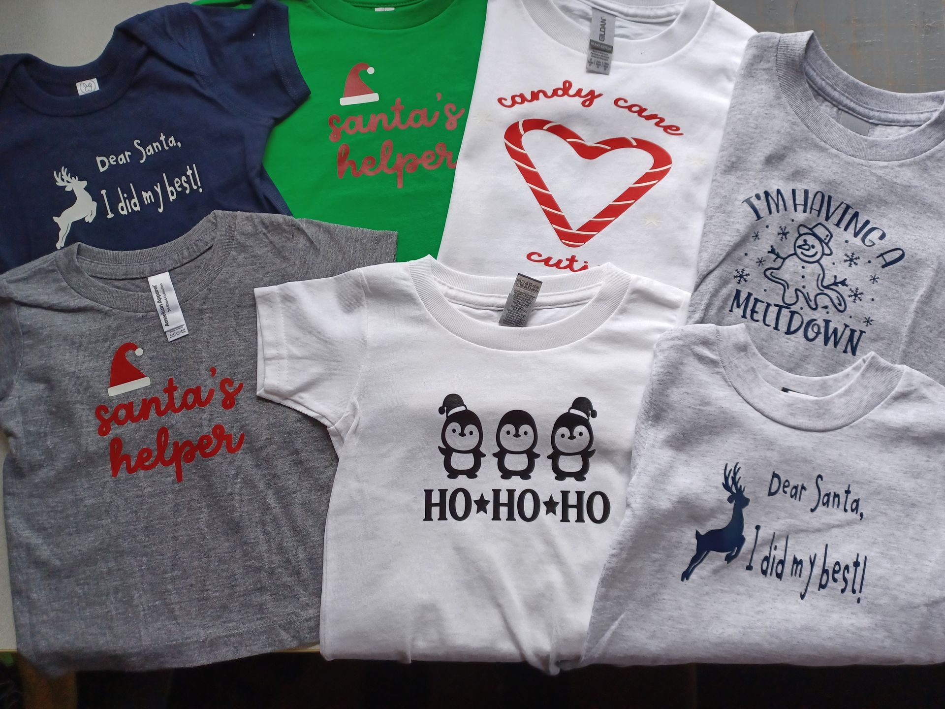 A collection of seven holiday-themed infant and children's shirts in green, gray, blue, and white with festive graphics.
