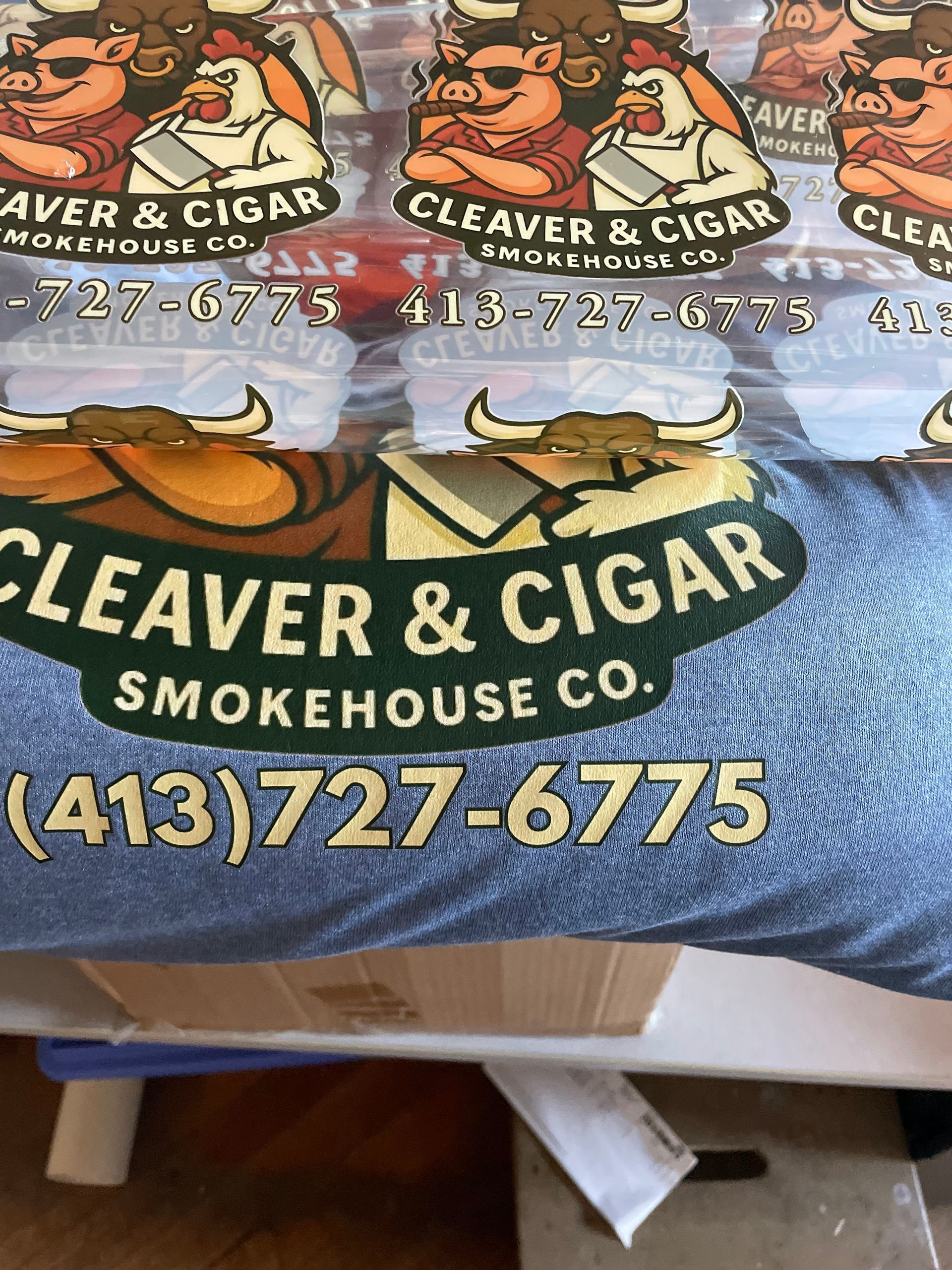 A logo for Cleaver & Cigar Smokehouse Co. featuring cartoon animals, with the phone number (413) 727-6775 printed below.