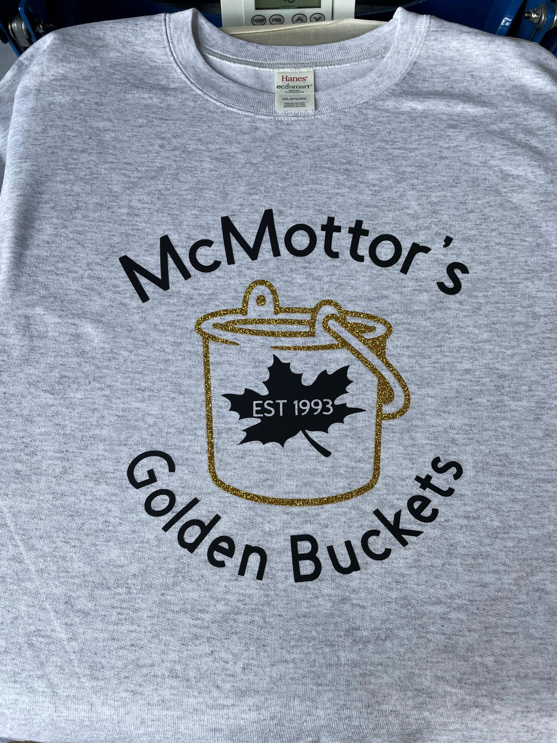 A grey t-shirt featuring a glittery gold bucket icon with a black maple leaf labeled 