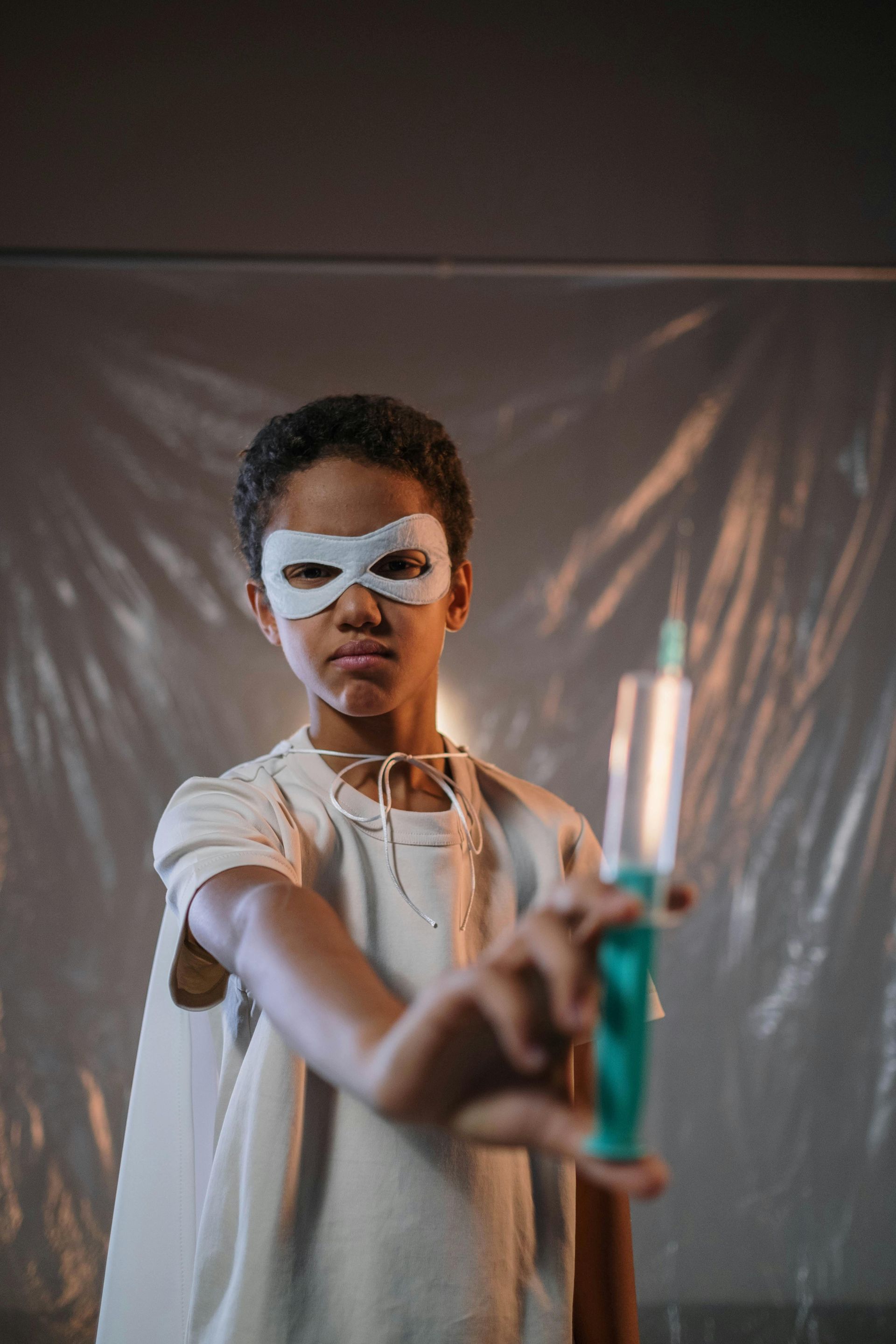 A young boy wearing a mask and cape is holding a syringe.