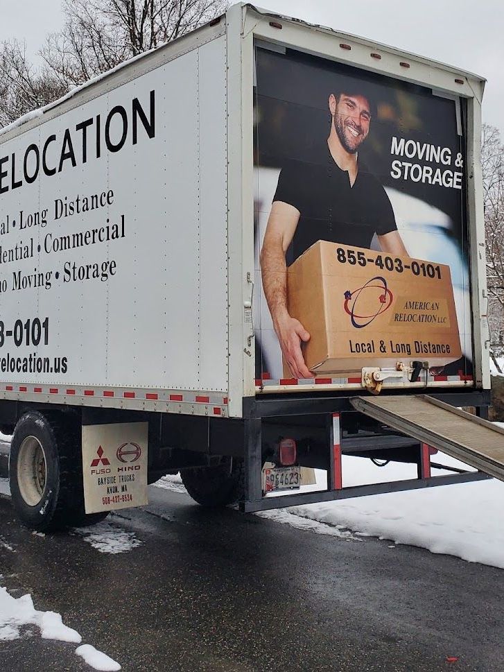 A white moving truck with a ramp down, parked in a snowy driveway. The truck has a large advertisement featuring a smiling man holding a box.