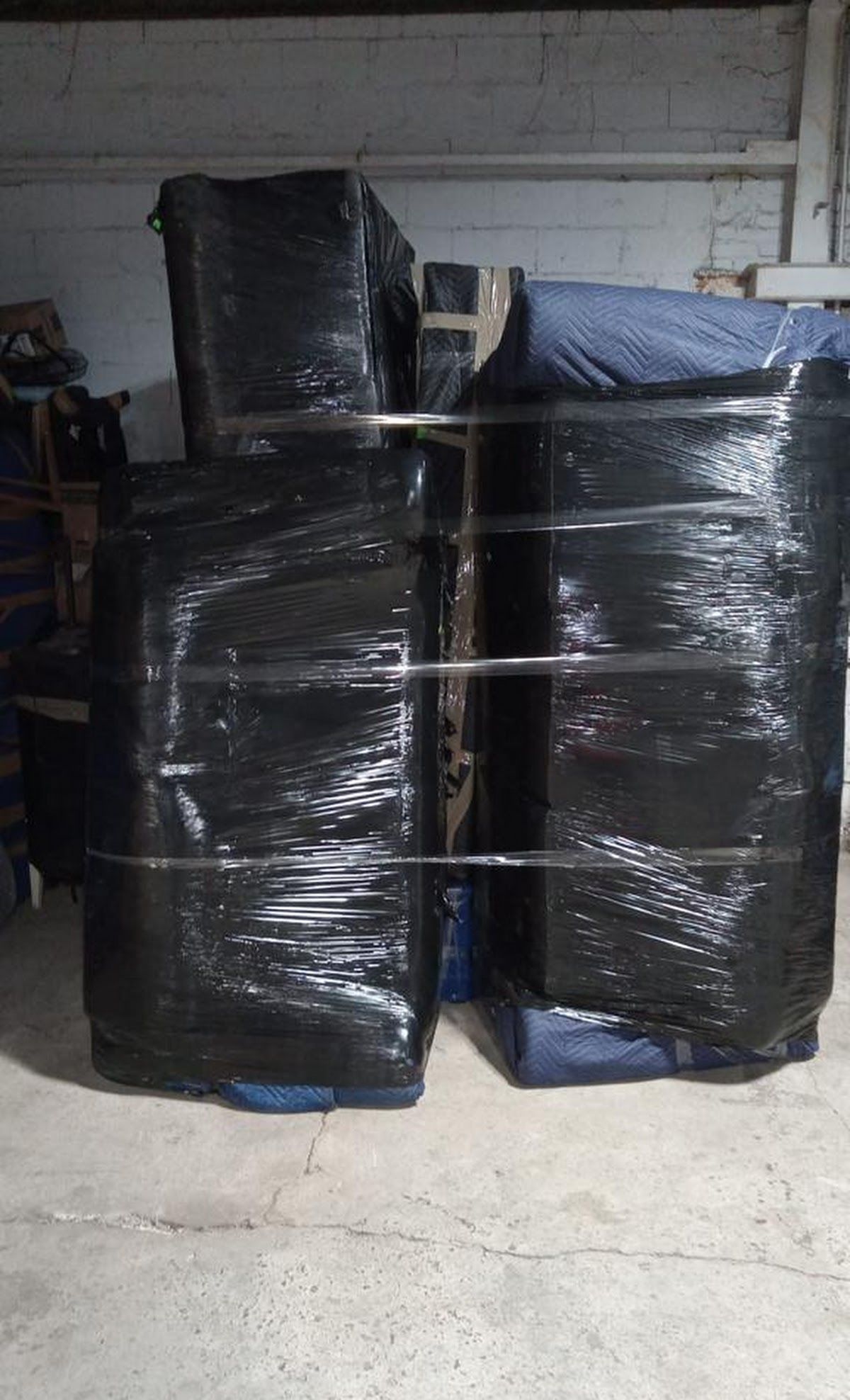 Several large furniture items, wrapped in black plastic, secured with straps, are stacked in a dimly lit storage space.