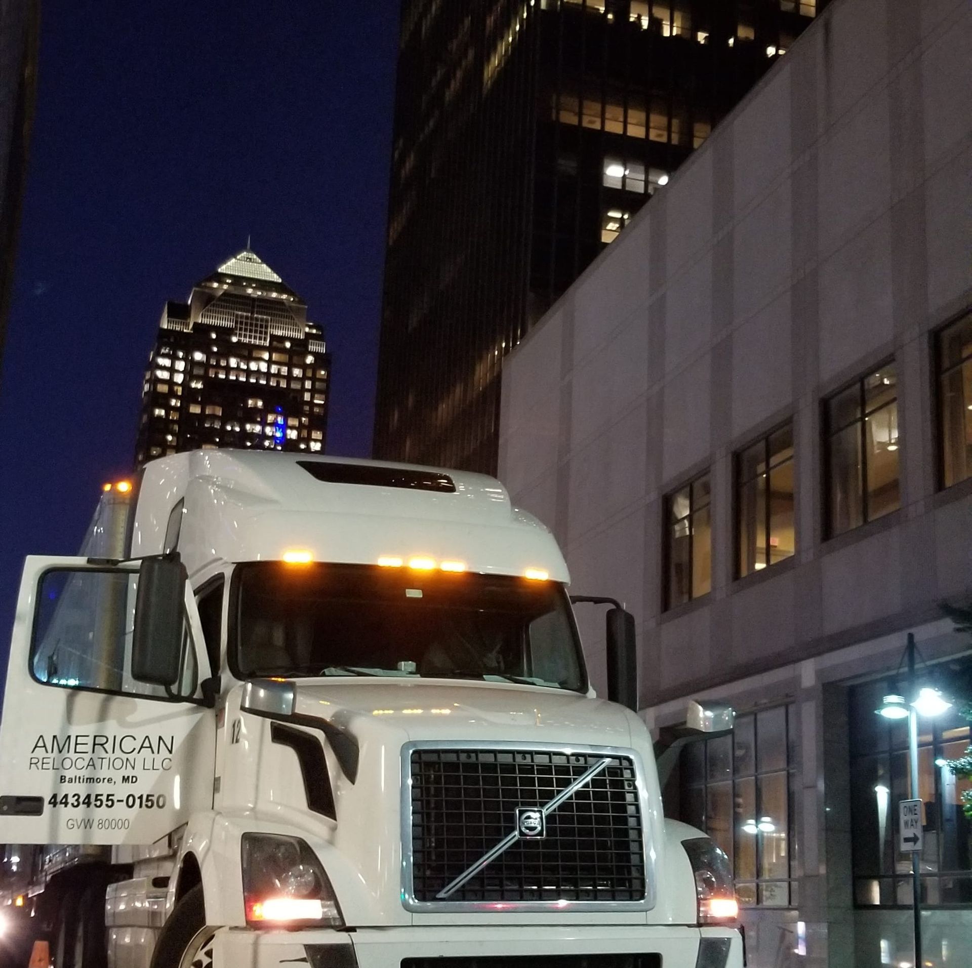 White semi-truck parked on a city street at night, doors open. Tall buildings and streetlights in the background.