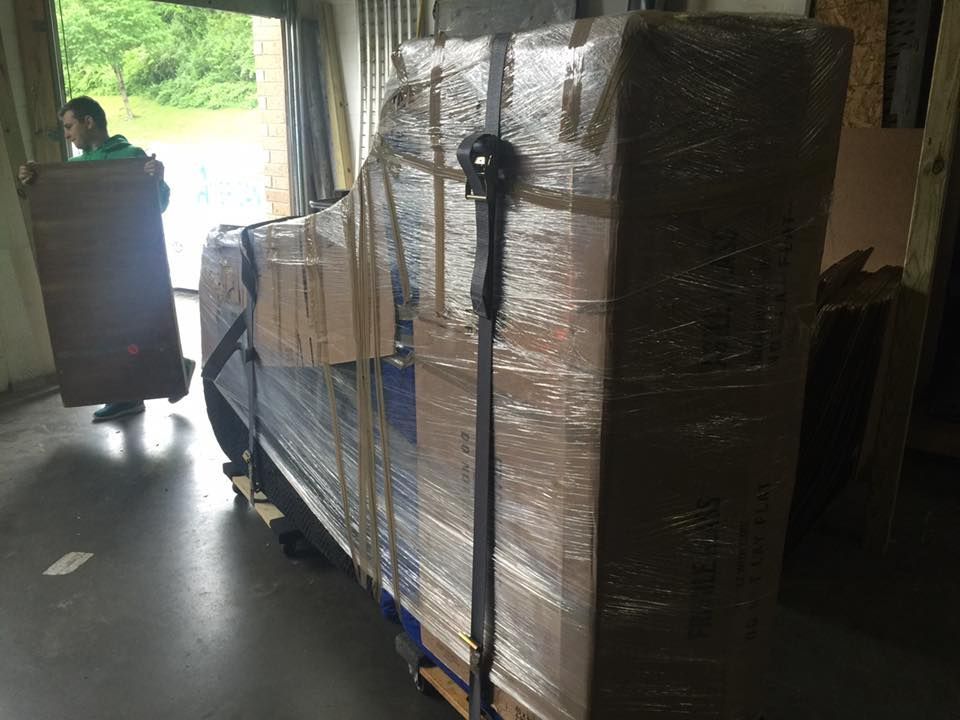 Piano wrapped in plastic and cardboard, secured with straps, being moved. A mover is in the background carrying a piece of furniture.