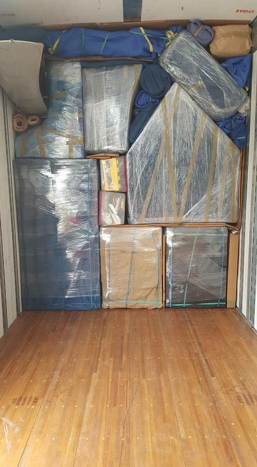 Packed moving truck interior filled with furniture and boxes wrapped in plastic and blue tarp, wooden floor.