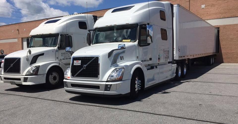 Two white semi-trucks parked on a paved area in front of a building. They are likely used for freight transport.
