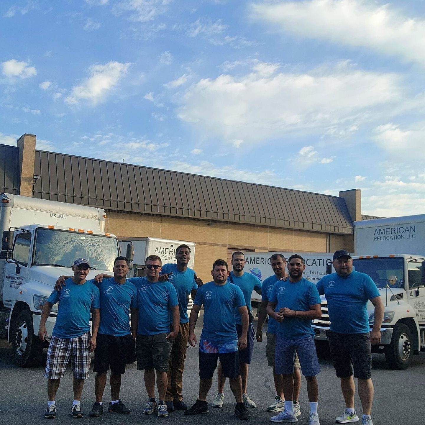 A group of movers in blue shirts and shorts stand in front of their moving trucks and a building.